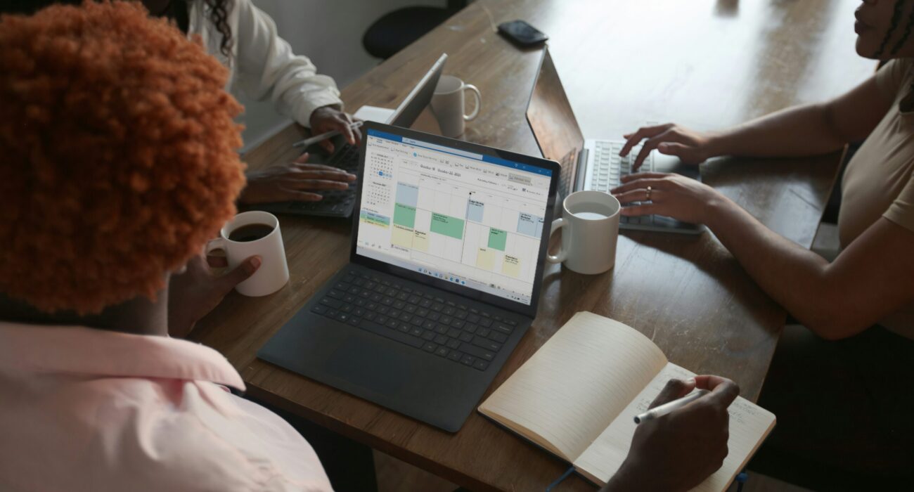 Three people in a meeting at a table discussing schedule on their Microsoft laptop